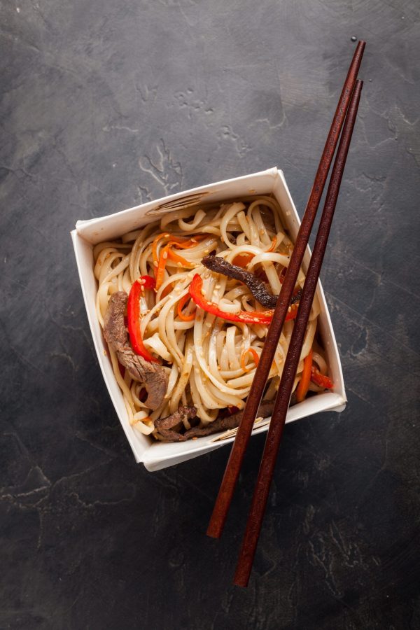 Close-up of noodles in a box with vegetables and beef in teriyaki sauce. Top view of Asian food, and wooden sticks. The concept of fast food.